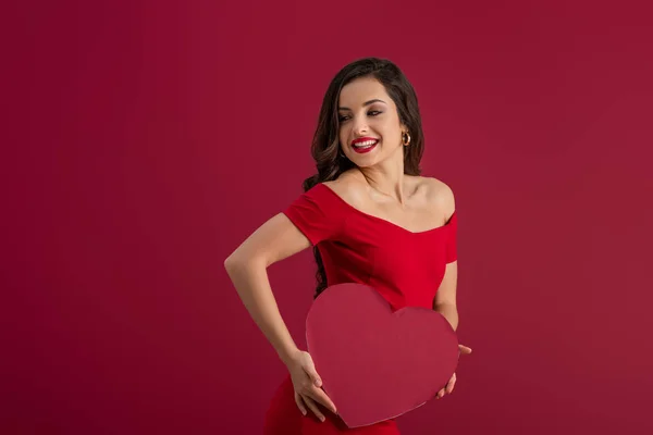 Сексуальная, элегантная девушка, держащая бумажное сердце и улыбающаяся изолированная на красном — стоковое фото