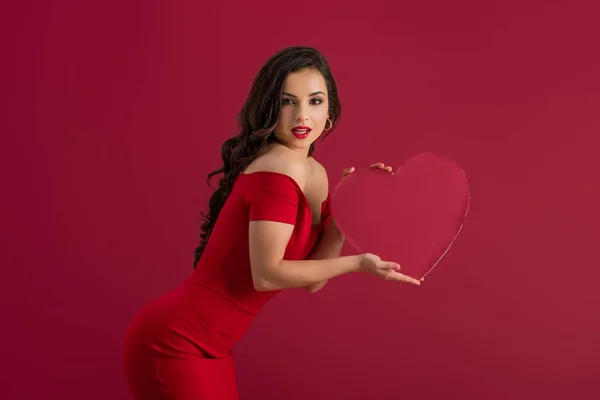 Seducente, ragazza elegante che tiene il cuore di carta e guardando la fotocamera isolata sul rosso — Foto stock