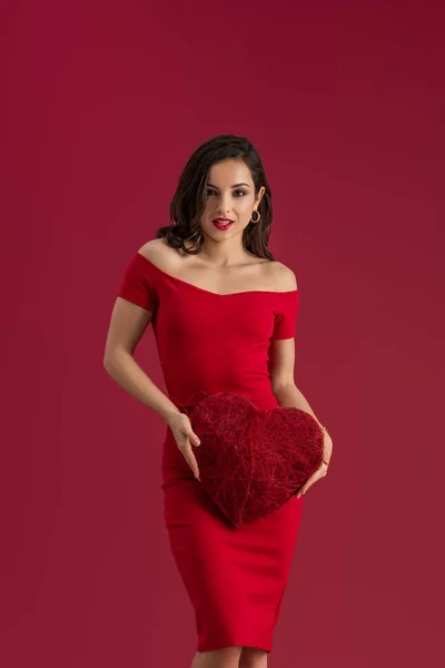 Sensual, elegant girl holding decorative heart and looking at camera isolated on red — Stock Photo