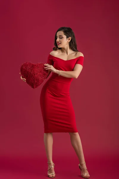 Sexy, elegante chica sosteniendo el corazón decorativo y sonriendo mientras está de pie sobre fondo rojo - foto de stock