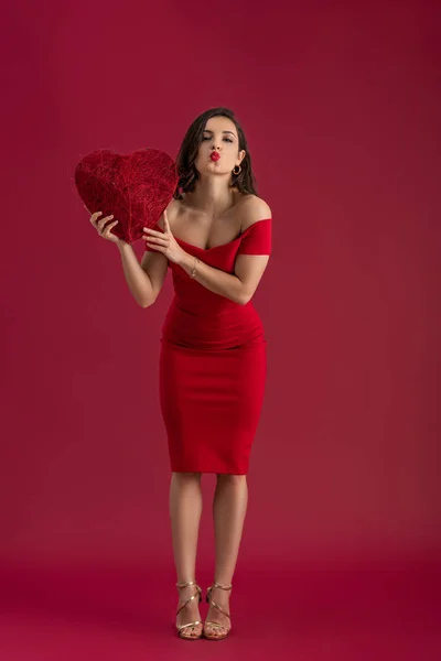 Coqueta, chica elegante sosteniendo el corazón decorativo y enviando un beso de aire a la cámara mientras está de pie sobre fondo rojo - foto de stock