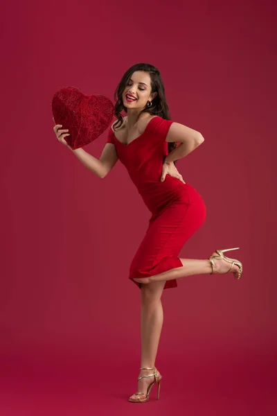 Niña sonriente y elegante sosteniendo el corazón decorativo mientras está de pie sobre una pierna en el fondo rojo - foto de stock