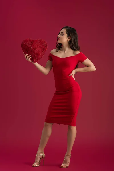 Sexy, menina elegante segurando coração decorativo enquanto de pé com a mão no quadril no fundo vermelho — Fotografia de Stock