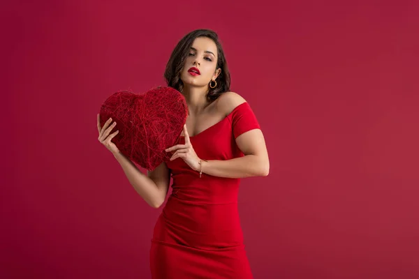 Seductive, elegant girl holding decorative heart while looking at camera isolated on red — Stock Photo