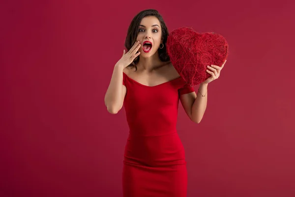 Surprised, elegant girl holding decorative heart and touching face while standing isolated on red — Stock Photo