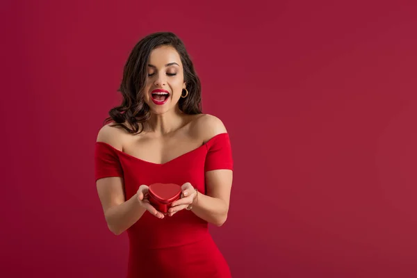 Emocionada, elegante chica sosteniendo caja de regalo en forma de corazón mientras está de pie aislado en rojo - foto de stock