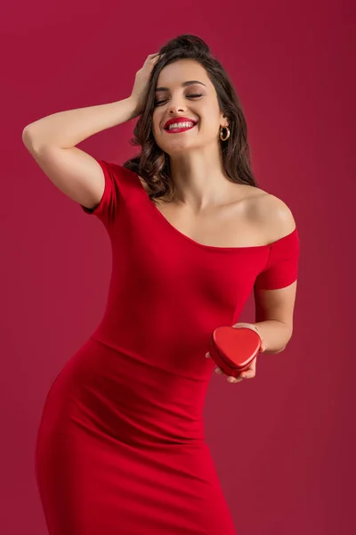 Щаслива, елегантна дівчина торкається волосся і тримає подарункову коробку у формі серця, стоячи з закритими очима ізольовано на червоному — стокове фото