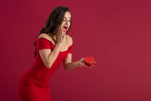 Sorprendida, elegante chica tocando la cara mientras sostiene la caja de regalo en forma de corazón aislado en rojo - foto de stock