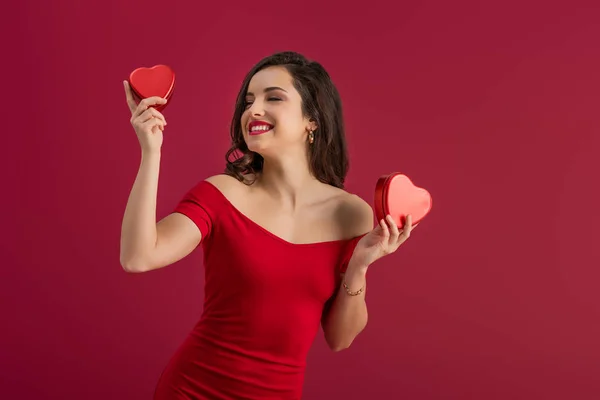 Chica sensual y elegante sosteniendo cajas de regalo en forma de corazón y sonriendo aislado en rojo - foto de stock