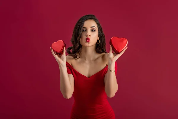 Sexy, elegante chica enviando aire beso en la cámara mientras sostiene en forma de corazón cajas de regalo aisladas en rojo - foto de stock