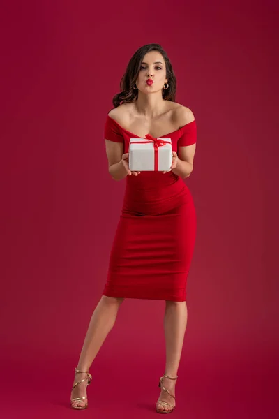 Сексуальна, елегантна дівчина тримає подарункову коробку і відправляє повітряний поцілунок на камеру на червоному тлі — стокове фото