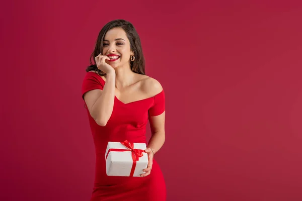 Sognante, elegante ragazza che tiene in mano la scatola regalo mentre distoglie lo sguardo e sorride isolato sul rosso — Foto stock