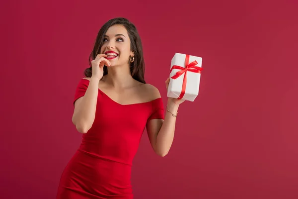 Sognante, elegante ragazza che tiene in mano la scatola regalo mentre distoglie lo sguardo e sorride isolato sul rosso — Foto stock