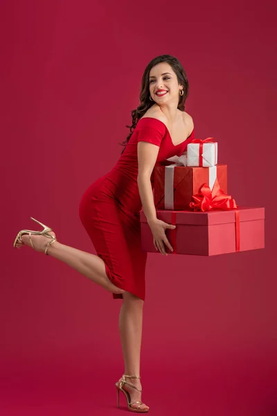 Кокетливая, элегантная девушка держит подарочные коробки и смотрит в сторону, стоя на одной ноге на красном фоне — стоковое фото