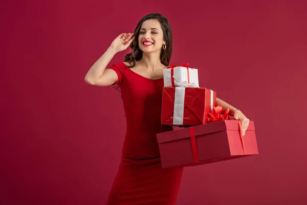 Sexy, chica feliz mirando hacia otro lado y agitando la mano mientras sostiene cajas de regalo aisladas en rojo - foto de stock