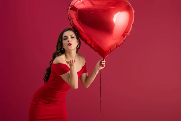 Seductora y elegante chica sosteniendo un globo en forma de corazón y enviando un beso de aire a la cámara aislada en rojo - foto de stock
