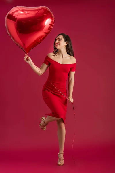 Glückliches, elegantes Mädchen mit herzförmigem Ballon auf einem Bein auf rotem Hintergrund stehend — Stockfoto