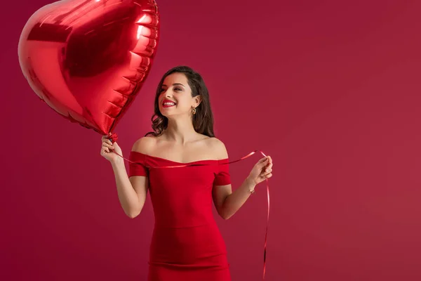 Heureuse, fille élégante regardant loin tout en tenant ballon en forme de coeur isolé sur rouge — Photo de stock
