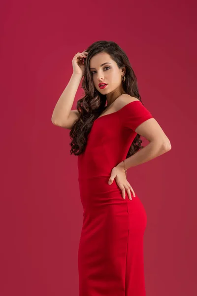 Sensual, elegante chica tocando el pelo mientras posa con la mano en la cadera aislado en rojo - foto de stock