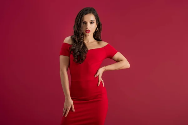 Menina sedutora em vestido elegante olhando para a câmera enquanto posando com a mão no quadril isolado no vermelho — Fotografia de Stock