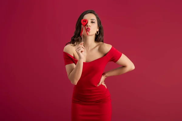 Sexy, elegante ragazza che copre l'occhio con il cuore giocattolo su bastone, mentre l'invio di aria bacio alla fotocamera isolata sul rosso — Foto stock