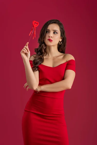 Мрійлива, елегантна дівчина дивиться геть, тримаючи іграшкове серце на паличці ізольовано на червоному — стокове фото