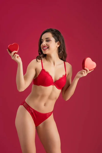Sexy, smiling girl in lingerie holding heart-shaped gift boxes isolated on red — Stock Photo