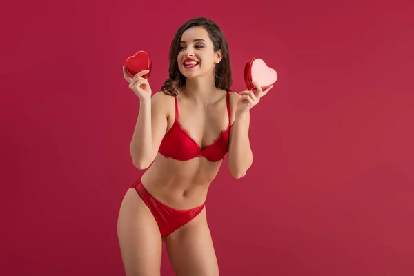 Sexy, smiling girl in lingerie looking away while holding heart-shaped gift boxes isolated on red — Stock Photo