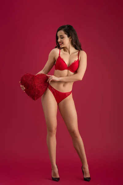 Seductive girl in lingerie and high heeled shoes holding decorative heart on red background — Stock Photo