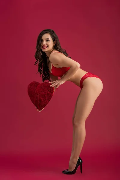 Chica sexy en lencería y zapatos de tacón alto sonriendo a la cámara mientras sostiene el corazón decorativo sobre fondo rojo - foto de stock
