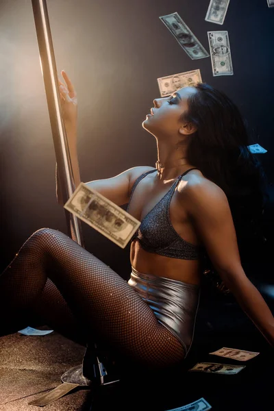 Side view of seductive stripper pole dancing near falling dollar banknotes on black — Stock Photo