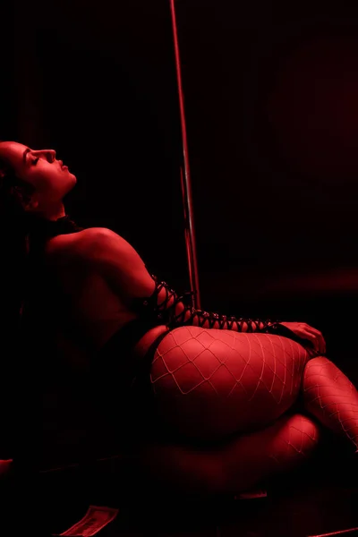 Iluminação vermelha em stripper apaixonado sentado perto de pilão em preto com iluminação vermelha — Fotografia de Stock