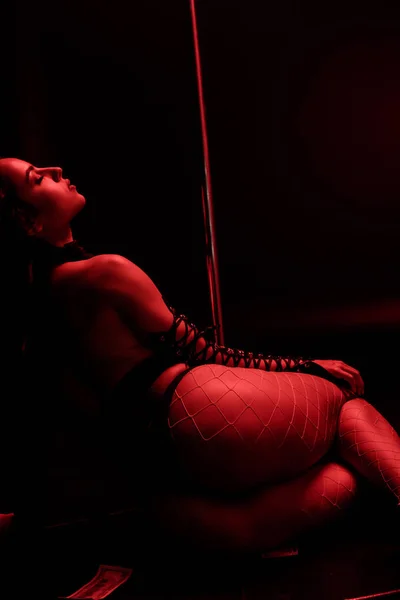 Red lighting on passionate stripper sitting near pylon on black with red lighting — Stock Photo