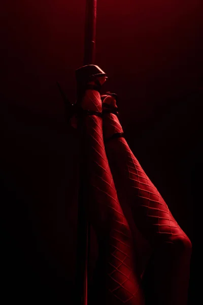 Cropped view of stripper in heels near pylon on black with red lighting — Stock Photo
