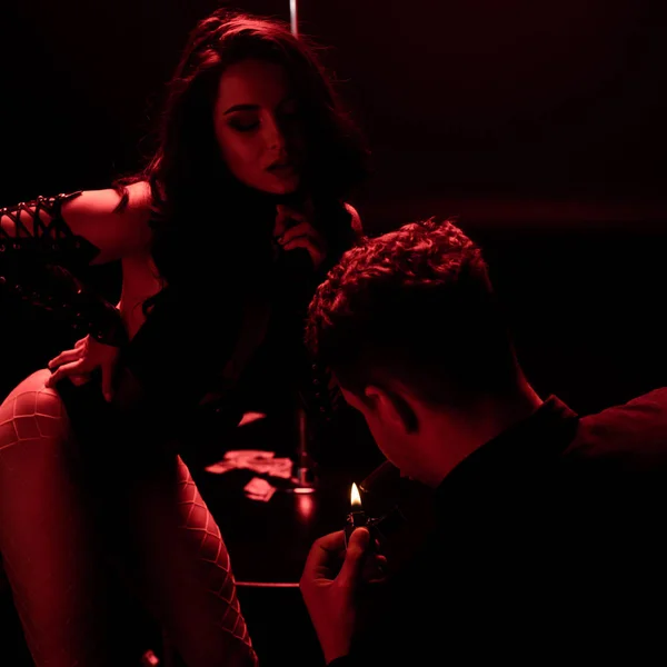 Man holding lighter while smoking near sexy stripper on black with red lighting — Stock Photo