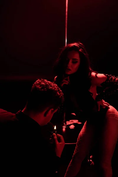 Hombre sosteniendo más ligero mientras fuma cerca de stripper seductora en negro con iluminación roja - foto de stock