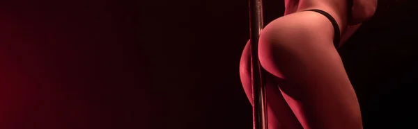 Panoramic shot of seductive stripper dancing near pylon on black with red lighting — Stock Photo