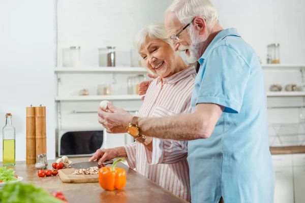 Vista lateral de la sonriente pareja de ancianos cocinando juntos en la mesa de la cocina - foto de stock