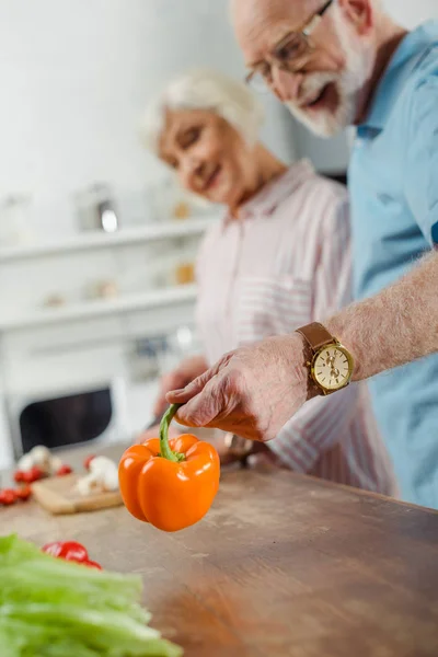 Enfoque selectivo del hombre mayor sosteniendo pimiento sonriendo esposa cortando verduras en la mesa de la cocina - foto de stock