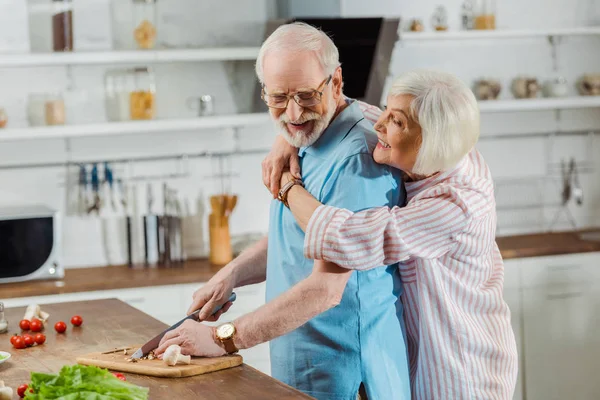 Vista lateral da mulher idosa abraçando o marido durante a cozinha na mesa da cozinha — Fotografia de Stock