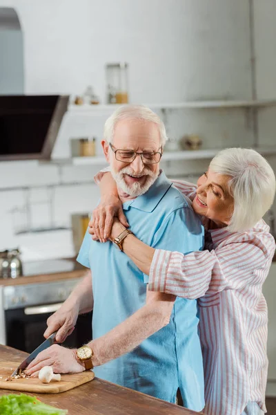 Vista lateral de la mujer sonriente abrazando al marido mientras corta verduras en la mesa de la cocina - foto de stock