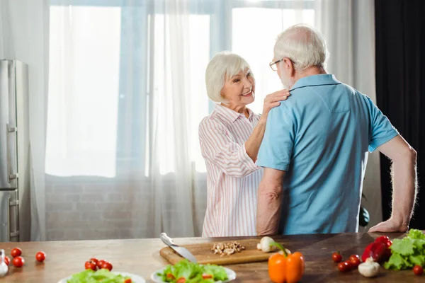 Enfoque selectivo de la mujer sonriente mirando al marido por verduras frescas en la mesa de la cocina - foto de stock