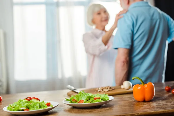 Enfoque selectivo de ensalada y verduras frescas en la mesa de la cocina y pareja de ancianos en segundo plano - foto de stock