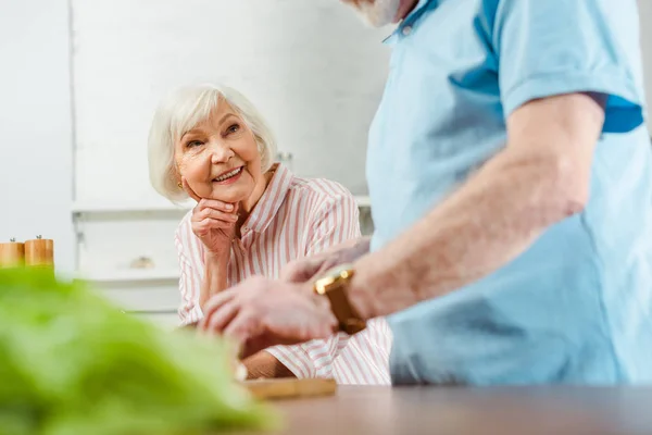 Выборочный фокус улыбающейся пожилой женщины, смотрящей на мужа во время приготовления пищи на кухонном столе — стоковое фото