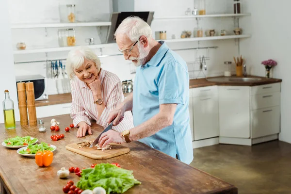 Улыбающаяся пожилая женщина, стоящая рядом с мужем, режет овощи на кухонном столе — стоковое фото