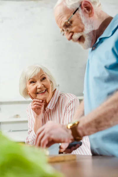 Selective focus of smiling woman looking at husband cutting vegetables on kitchen table — Stock Photo