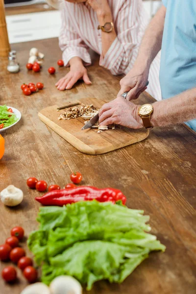 Vista cortada do homem sênior cortando cogumelos enquanto cozinha por esposa na mesa da cozinha — Fotografia de Stock