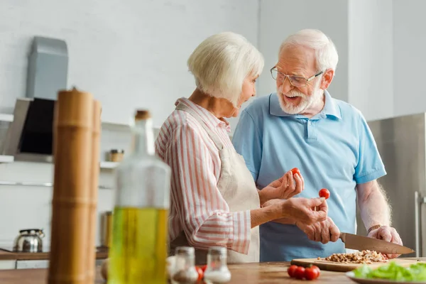 Selective focus of smiling senior man looking at wife while cutting vegetables on kitchen table — Stock Photo