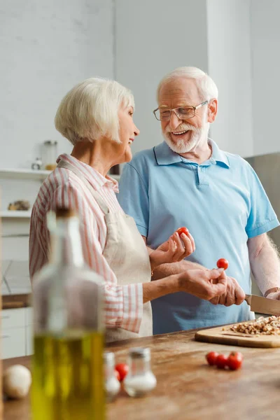 Selektiver Fokus eines lächelnden Mannes, der seine Frau mit Kirschtomaten anschaut, während er Pilze auf dem Küchentisch schneidet — Stockfoto