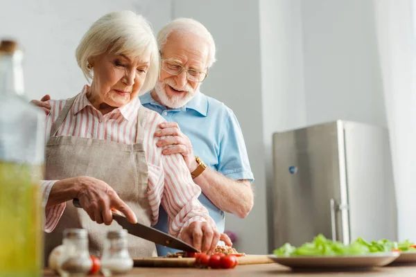 Выборочный фокус улыбающегося пожилого мужчины, обнимающего жену во время приготовления пищи на кухонном столе — стоковое фото